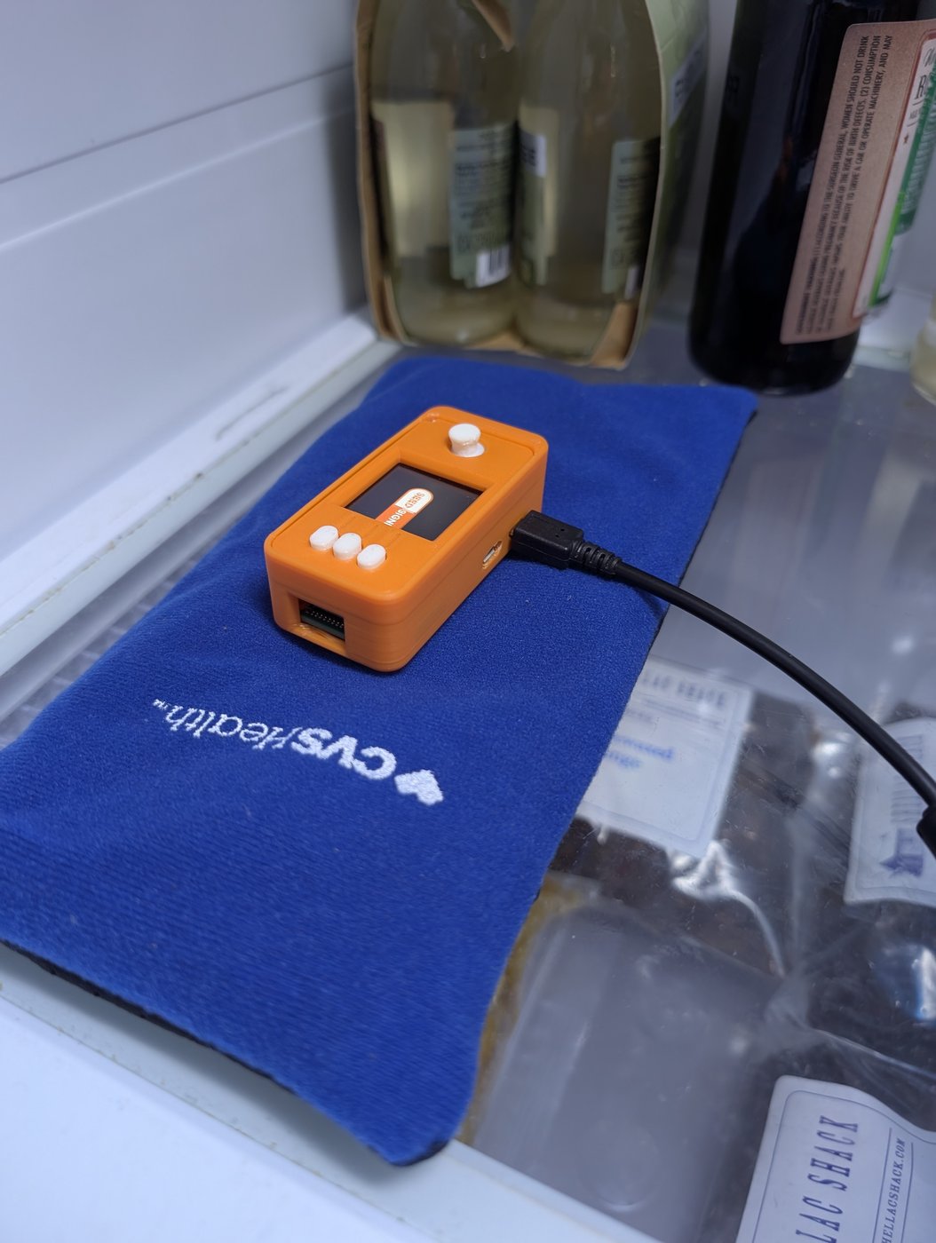 A SeedSigner in an orange case resting camera-down on a blue gel pack in its fabric sleeve on a refrigerator shelf, screen lit, USB cable running off-frame.
