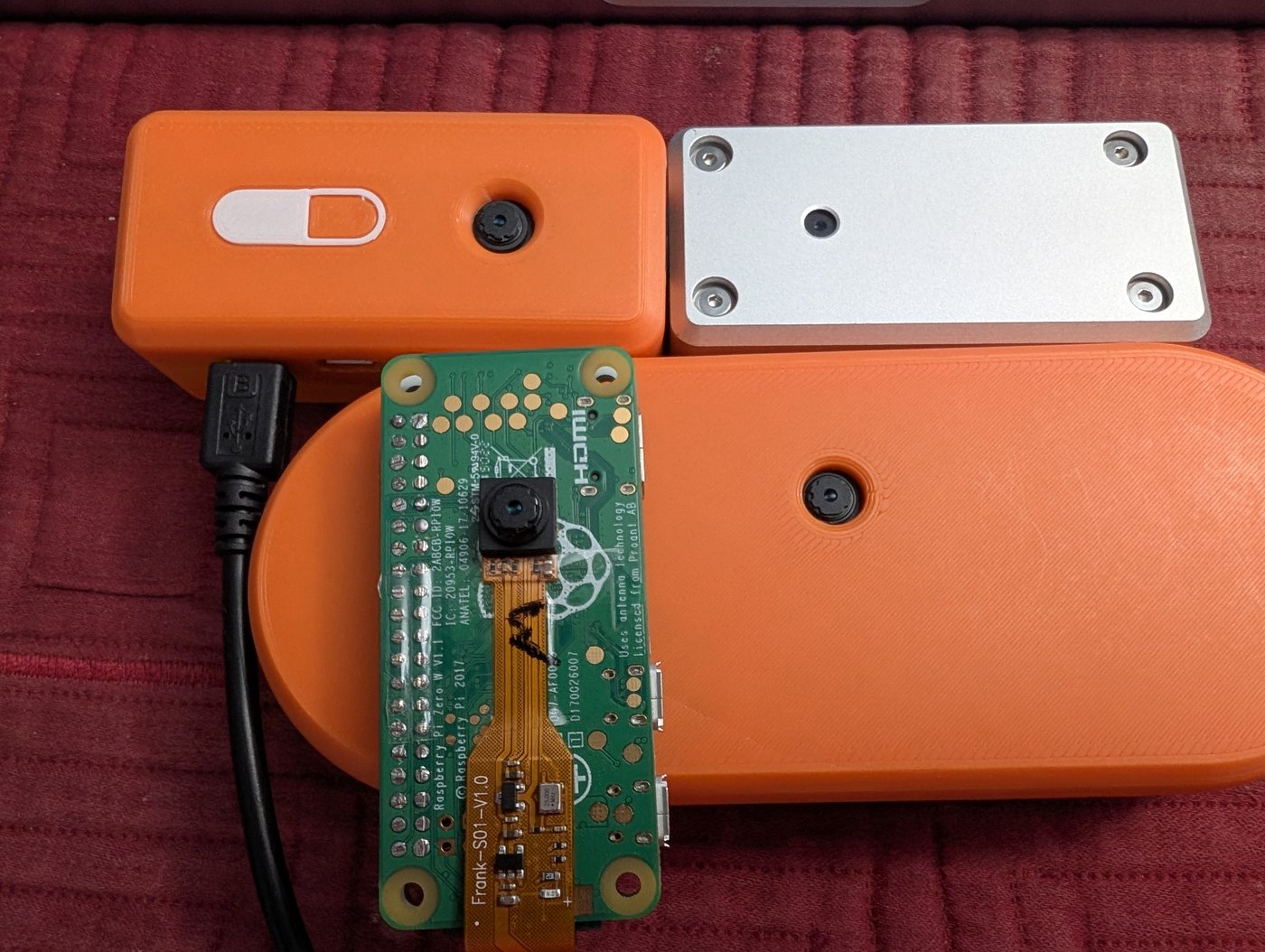 Four SeedSigner units camera-side up: an orange 3D-printed case, a milled aluminum case, a bare Pi Zero board with its camera ribbon taped on, and a large oval Plus-style case.